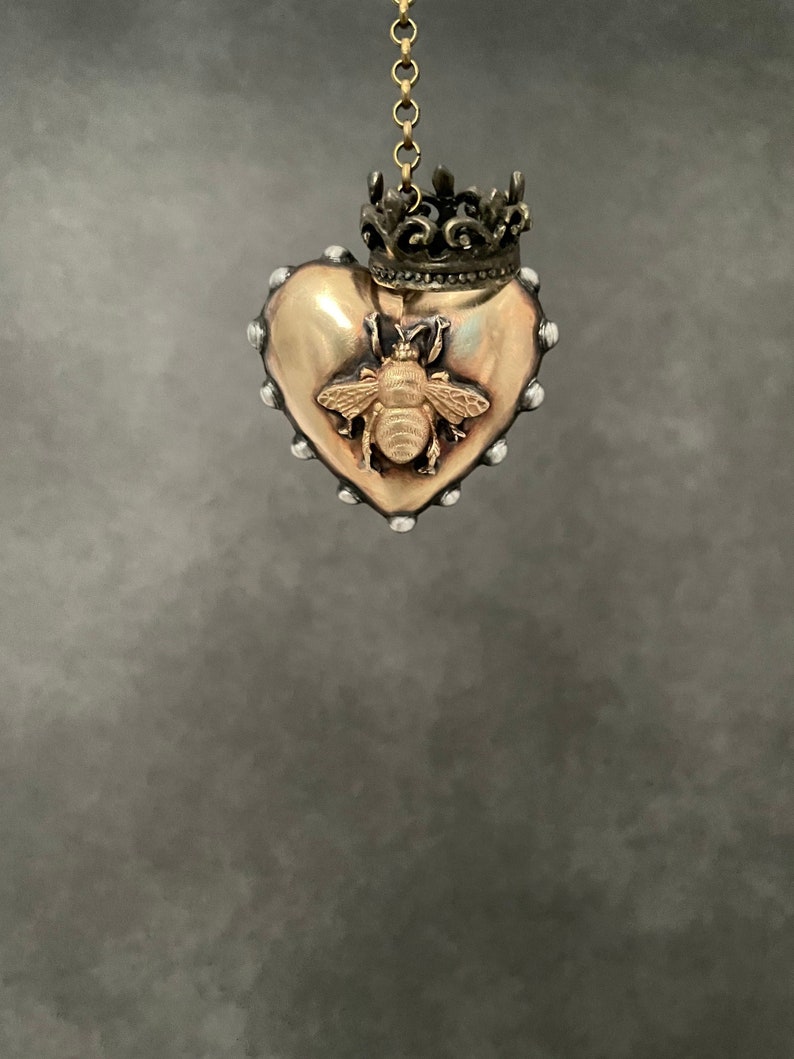 Puede incluir: Un colgante de coraz&oacute;n dorado con un dise&ntilde;o de abeja y una peque&ntilde;a corona en la parte superior. El colgante est&aacute; colgado de una cadena.