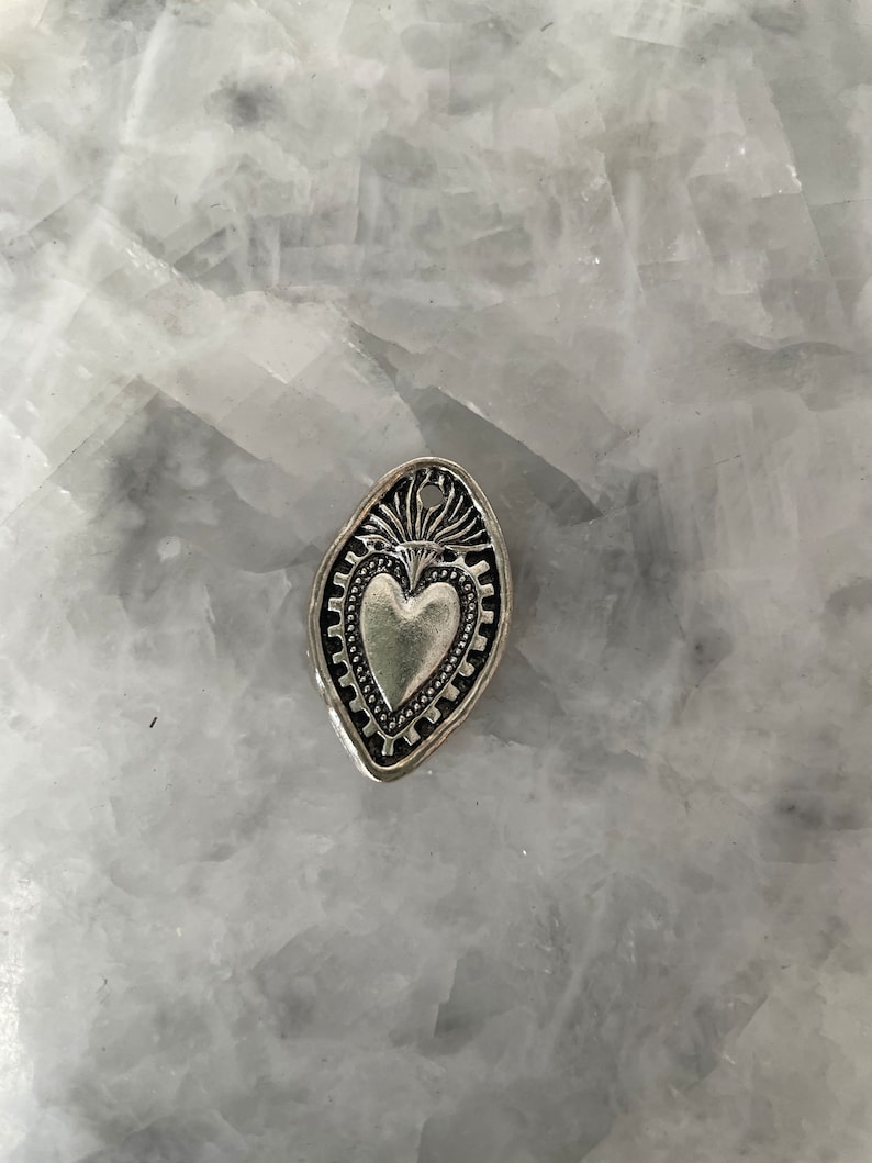 May include: A silver charm in the shape of a heart with a decorative border. The heart is raised and has a textured surface.