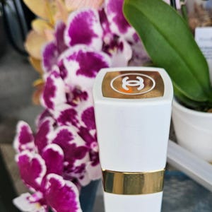 Puede incluir: Un tubo de pintalabios blanco con una banda dorada en el centro y una tapa dorada que presenta el logotipo de Chanel en blanco. El envase tiene una forma rectangular con bordes ligeramente redondeados y un acabado liso y brillante.
