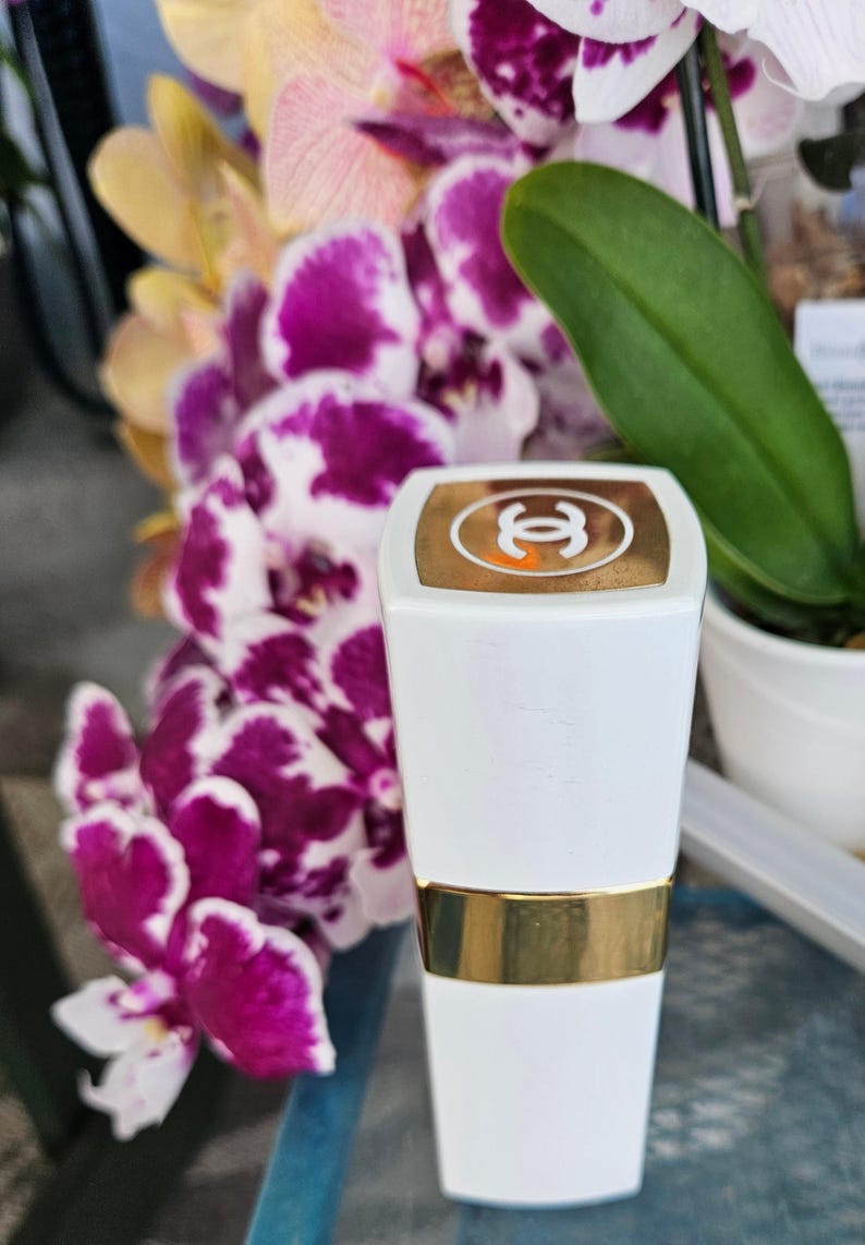 Puede incluir: Un tubo de pintalabios blanco con una banda dorada en el centro y una tapa dorada que presenta el logotipo de Chanel en blanco. El envase tiene una forma rectangular con bordes ligeramente redondeados y un acabado liso y brillante.