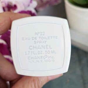 Puede incluir: Una base de frasco de perfume rectangular blanca con texto en relieve que dice N&deg;22 EAU DE TOILETTE SPRAY CHANEL 1.7 FL. OZ. 50 ML CHANEL Inc. N.Y., N.Y. 10019. El objeto presenta un acabado liso y mate con un borde elevado alrededor del texto.