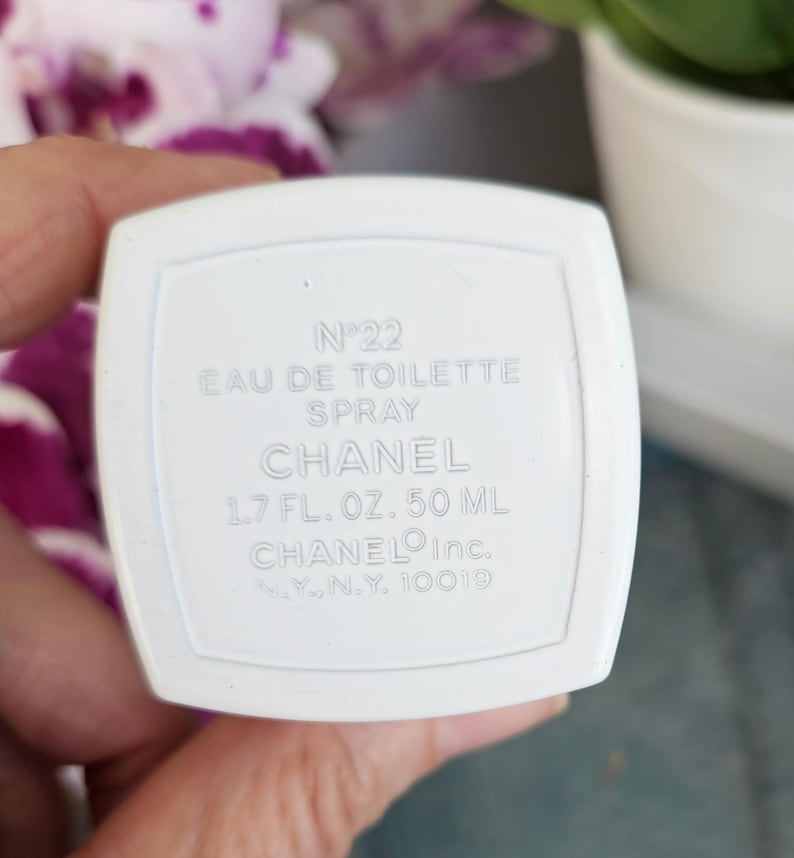 Puede incluir: Una base de frasco de perfume rectangular blanca con texto en relieve que dice N&deg;22 EAU DE TOILETTE SPRAY CHANEL 1.7 FL. OZ. 50 ML CHANEL Inc. N.Y., N.Y. 10019. El objeto presenta un acabado liso y mate con un borde elevado alrededor del texto.