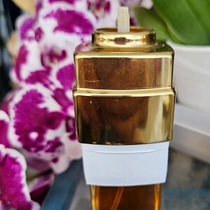Puede incluir: Un frasco de perfume rectangular con una base de vidrio transparente que contiene l&iacute;quido &aacute;mbar, una banda blanca alrededor del centro y una tapa met&aacute;lica dorada reflectante. Una peque&ntilde;a boquilla de pl&aacute;stico sobresale de la parte superior.
