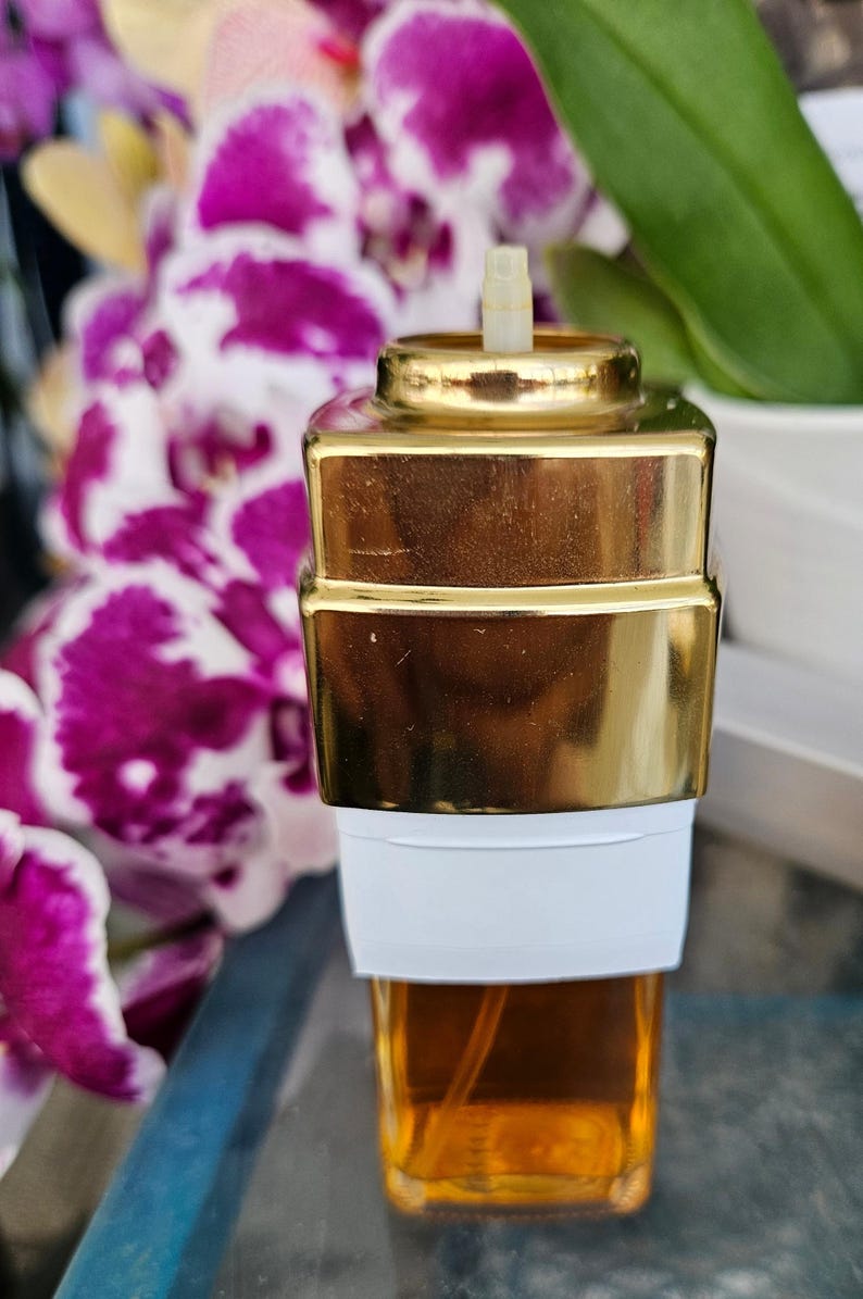Puede incluir: Un frasco de perfume rectangular con una base de vidrio transparente que contiene l&iacute;quido &aacute;mbar, una banda blanca alrededor del centro y una tapa met&aacute;lica dorada reflectante. Una peque&ntilde;a boquilla de pl&aacute;stico sobresale de la parte superior.