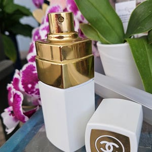 Puede incluir: Un frasco de perfume rectangular blanco con un atomizador met&aacute;lico dorado y una tapa blanca a juego que presenta una parte superior cuadrada dorada con el logotipo CC entrelazado en blanco. El frasco tiene un acabado liso y dise&ntilde;o geom&eacute;trico.