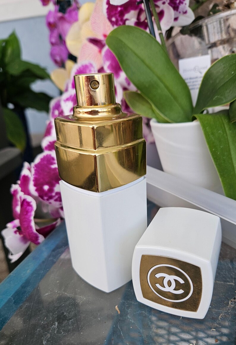 Puede incluir: Un frasco de perfume rectangular blanco con un atomizador met&aacute;lico dorado y una tapa blanca a juego que presenta una parte superior cuadrada dorada con el logotipo CC entrelazado en blanco. El frasco tiene un acabado liso y dise&ntilde;o geom&eacute;trico.