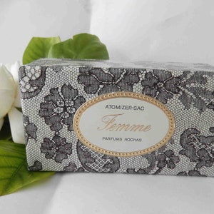 May include: A vintage perfume box with a black lace pattern on a white background. The box has a gold-trimmed oval label with the word "Femme" in elegant script. A white flower and green leaves are in the background.