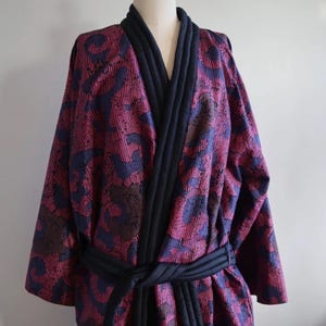 Könnte beinhalten: Gemusterter Bademantel in Rot-, Lila- und Marineblautönen, auf einer Schaufensterpuppe präsentiert. Der Bademantel hat ein geometrisches Design und eine marineblaue Steppkante am Kragen und an den Kanten. Ein passender marineblauer Gürtel ist um die Taille gebunden.