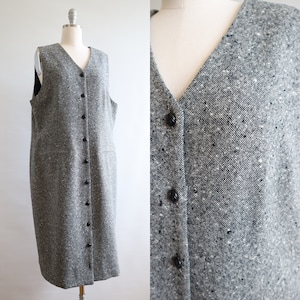 May include: A grey tweed dress with a V-neck and a button-down front. The sleeveless dress has two front pockets and is displayed on a mannequin. The fabric has flecks of white and black.