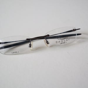 May include: Rimless Oakley spectacles with clear lenses and a dark metal frame. The brand name "OAKLEY" and "LENS J" are printed on the lens. The glasses have a minimalist design.