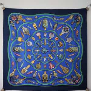 May include: A square silk scarf with a navy blue border and a central design of ornate perfume bottles in various shapes and colors. The bottles are arranged in concentric circles on a royal blue background, with green accents.