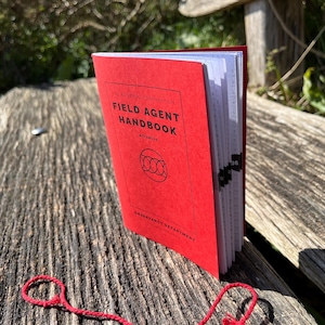 Puede incluir: Un libro rojo cerrado con el título "Field Agent Handbook" en la portada. El libro está sobre una superficie de madera con un lazo de cuerda rojo cerca.