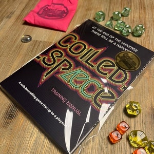 Könnte beinhalten: Eine schwarz-weiße Spielbox mit dem Titel "COILED SPACE" in einer Retro-Schriftart. Die Box ist in Plastik verpackt und hat ein Etikett mit der Aufschrift "Training Manual". Das Spiel wird als Solo-Tagebuchspiel für bis zu 4 Spieler beschrieben. Auf einer Holzoberfläche liegen mehrere grüne und gelbe Würfel.
