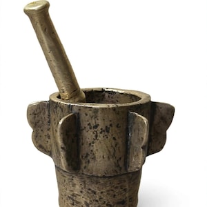 May include: A heavy, dark metallic mortar and pestle set with a weathered, mottled finish. The mortar features a cylindrical body with four protruding, wing-shaped vertical flanges around the exterior, and a matching pestle rests inside.