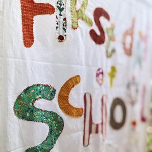 Puede incluir: Una pancarta de tela blanca con las palabras First Day of School aplicadas en varias telas coloridas con patrones. Cada letra está cosida individualmente sobre el fondo blanco con hilo visible, creando una apariencia texturizada y superpuesta.