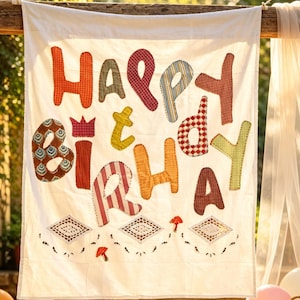 May include: A white fabric banner featuring the text HAPPY BIRTHDAY in multicolored, patterned fabric letters. The design includes a small red crown icon above the letter I, and three white lace diamond shapes with two small red mushroom icons.