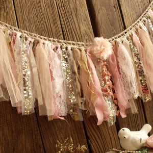 May include: A delicate, handmade fabric garland with pink, beige, and gold accents. The garland is made of various textures, including lace, tulle, and sequins. It is perfect for adding a touch of whimsy and elegance to any event.