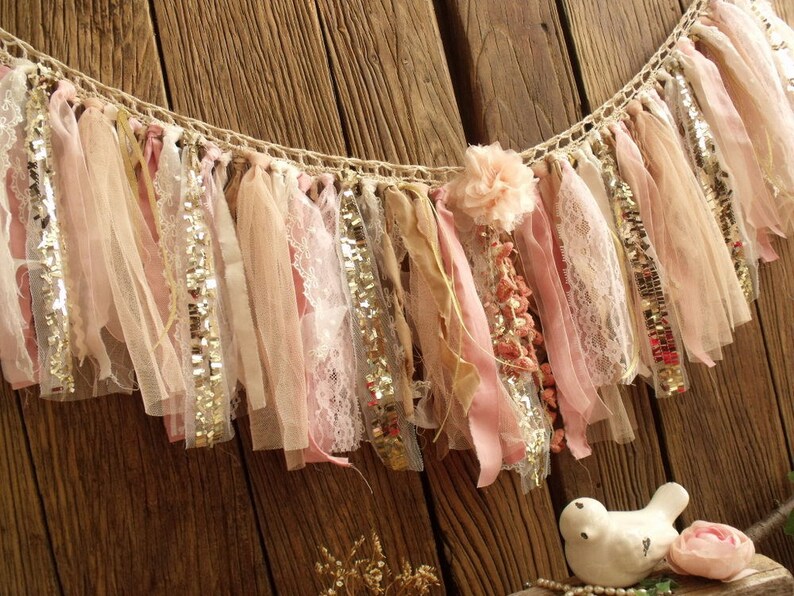 May include: A delicate, handmade fabric garland with pink, beige, and gold accents. The garland is made of various textures, including lace, tulle, and sequins. It is perfect for adding a touch of whimsy and elegance to any event.