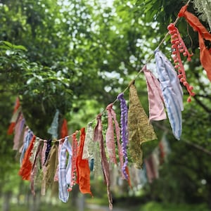 Pode incluir: Uma guirlanda decorativa feita de várias tiras de tecido amarradas a um cordão. As tiras apresentam uma mistura de texturas e cores, incluindo laranja, rosa, lavanda, verde-oliva e padrões em azul e branco, com algumas peças contendo renda.