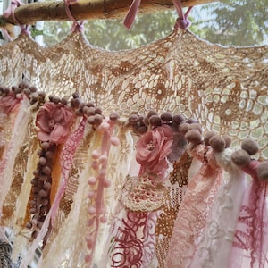 May include: A decorative, handmade curtain featuring a natural wood rod and a crocheted top. The curtain is adorned with various fabrics in shades of cream, pink, and brown, including lace, ribbons, and floral accents. Small pom-poms add texture.