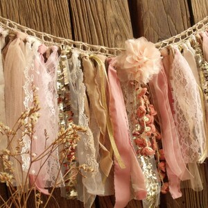 May include: A delicate, handmade banner with pink, beige, and gold fabric strips, lace, and sequins. The banner is strung on a crocheted rope and features a pink fabric flower.