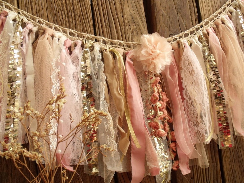 May include: A delicate, handmade banner with pink, beige, and gold fabric strips, lace, and sequins. The banner is strung on a crocheted rope and features a pink fabric flower.