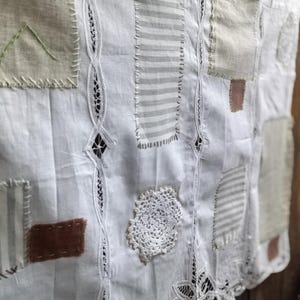May include: A white textile wall hanging featuring vertical strips of intricate lace insertion. The fabric is decorated with various rectangular patches in beige, grey stripes, and brown, along with a circular white crochet doily appliqué.