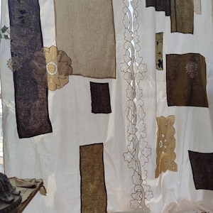 Könnte beinhalten: Cremefarbener Vorhang mit rechteckigen Flecken in Braun- und Olivgrüntönen. Ein dekoratives Blumenmuster verläuft vertikal in der Mitte, und eine große Blumenapplikation befindet sich auf der linken Seite. Der Vorhang hat eine strukturierte Optik.