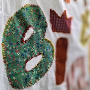 May include: A white fabric banner featuring the letter B appliqued in a green floral print fabric with yellow and red hand-stitched edges. Nearby, a dark red checkered crown shape and other fabric letters are visible on the white background.