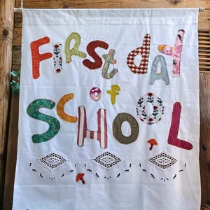 Könnte beinhalten: Ein weißer Wandbehang aus Stoff mit dem applizierten Text "First day of SCHOOL" aus verschiedenen bunten Stoffen mit Mustern. Das Banner enthält drei weiße, gehäkelte, rautenförmige Spitzenakzente und kleine gestickte Pilzmotive.
