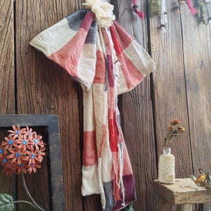 Puede incluir: Una bufanda estampada en tonos rojos, blancos y azules cuelga sobre una pared de madera rústica. La bufanda está adornada con una flor blanca y tiene bordes deshilachados. Los elementos decorativos incluyen una flor de metal enmarcada y un pequeño taburete de madera.