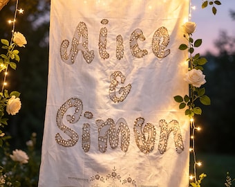 May include: A rectangular white fabric banner featuring the embroidered text Alice & Simon in a decorative lace-like font. The banner has a scalloped lace trim at the bottom and is illuminated by warm string lights draped around it.
