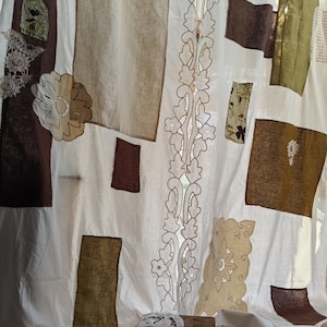 Puede incluir: Cortina blanca con parches de tela marrón, verde oliva y beige. La cortina presenta un diseño vertical central con recortes florales y elementos de encaje decorativos. Un pequeño taburete de madera sostiene el borde inferior de la cortina.