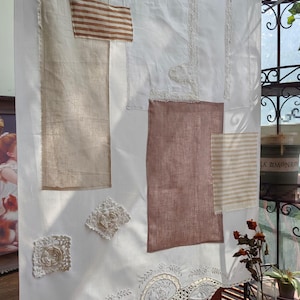 May include: White curtain with various fabric patches in beige, brown, and striped patterns. Decorative lace and crochet details are sewn onto the curtain. The curtain is hanging in front of a window with a wrought iron railing.