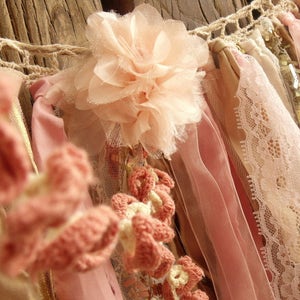 May include: A pink and white fabric garland with crocheted flowers, lace, and ribbons. The garland is made of various textures and materials, creating a delicate and feminine look.