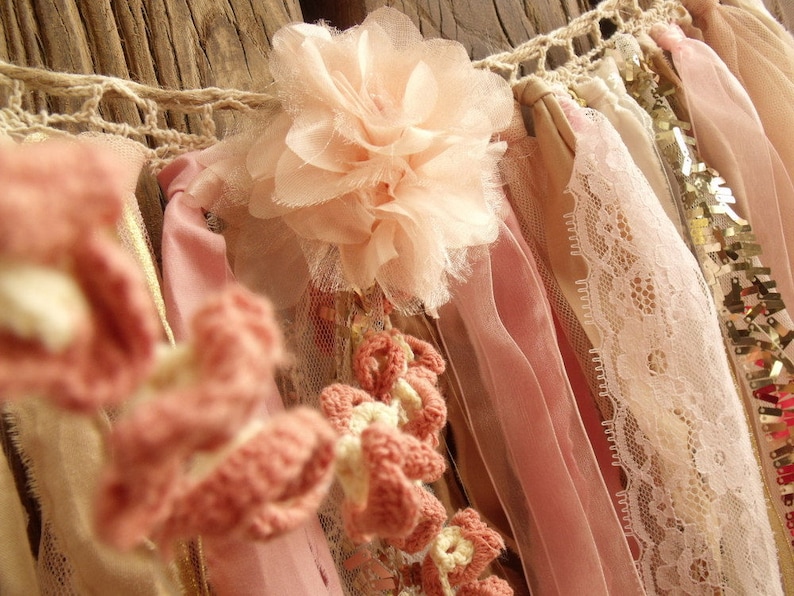 May include: A pink and white fabric garland with crocheted flowers, lace, and ribbons. The garland is made of various textures and materials, creating a delicate and feminine look.