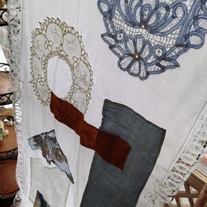 May include: White fabric with appliqued lace and fabric patches. Features a large blue lace doily, a gold lace circle, and a brown fabric accent. The edges are trimmed with white lace. A unique textile art piece.