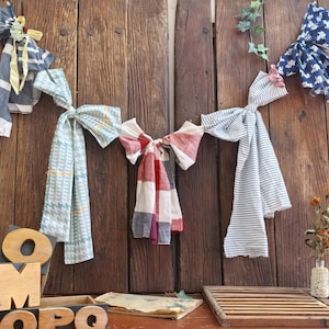 Puede incluir: Surtido de lazos de tela en varios patrones y colores, incluyendo rayas azules y blancas, cuadros y diseños florales. Los lazos cuelgan de una puerta de madera rústica. Elementos decorativos incluyen flores secas y bloques de letras de madera.