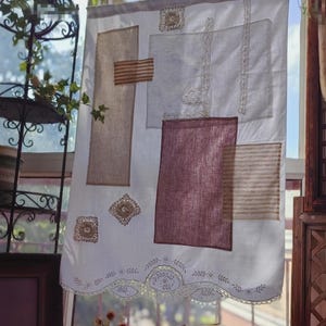 Puede incluir: Cortina blanca con parches de tela en tonos beige y burdeos. La cortina tiene detalles bordados y un borde inferior festoneado. Los parches son rectangulares y cuadrados, algunos con patrones de rayas.