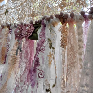 May include: A selection of lace curtains in shades of white, pink, and beige. The curtains are decorated with floral details and hang from a rod with pom-pom decorations. The style is vintage and romantic, with a delicate, handcrafted feel.