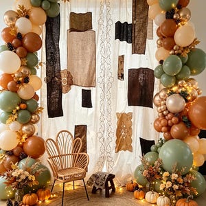 May include: A decorative event backdrop featuring two large balloon arches in shades of sage green, cream, tan, and metallic gold, accented with pinecones and autumn leaves. A rattan chair and small wooden stool sit on a round jute rug.