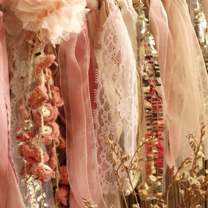 May include: A pink and cream fabric garland with crocheted flowers, lace, and sequins. The garland is hanging from a wooden surface.