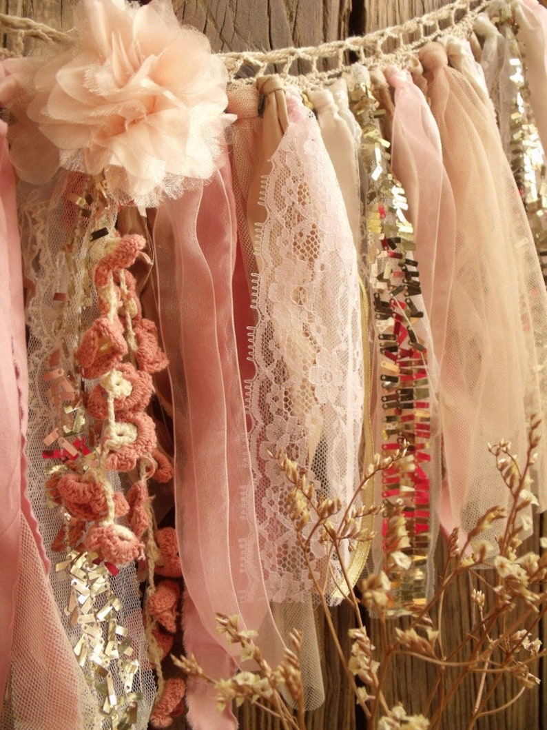 May include: A pink and cream fabric garland with crocheted flowers, lace, and sequins. The garland is hanging from a wooden surface.