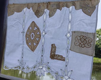 Könnte beinhalten: Weißer Vorhang mit aufwendiger brauner und beiger Stickerei. Der Vorhang zeigt florale und geometrische Muster, darunter eine große zentrale Raute und eine dekorative Bordüre. Der obere Rand hat einen gewellten Besatz. Der Vorhang hängt vor einem Gewässer.