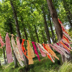 Puede incluir: Una guirnalda decorativa que presenta una serie de tiras de tela colgantes en varios tonos de rosa, naranja y rojo. Las tiras están atadas a una cuerda central e incluyen una mezcla de colores sólidos, patrones florales y telas texturizadas.