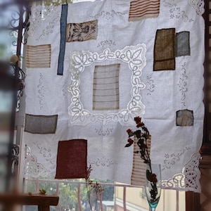 Może przedstawiać: Biała lniana zasłona z haftowanymi kwiatowymi detalami i łatkami z różnych tkanin. Zasłona posiada duży, ozdobny kwadrat z koronki pośrodku i koronkowe wykończenie na krawędziach. Z przodu szklanka z suszonymi kwiatami.