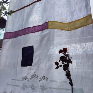 Pode incluir: Cortina de linho branco com faixas horizontais coloridas: azul-petróleo, castanho, roxo e amarelo. Um quadrado roxo escuro é costurado no tecido. A borda inferior apresenta padrões florais e geométricos bordados. Um vaso contém rosas vermelhas secas.