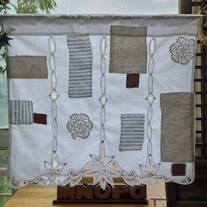 Puede incluir: Un panel de tela blanca con inserciones de encaje verticales y un borde de encaje decorativo en la parte inferior. El panel está adornado con varios parches rectangulares de tela beige, a rayas grises y marrón, junto con dos tapetes de encaje.