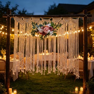 May include: A cream-coloured macrame wall hanging suspended from a dark wooden frame, featuring intricate knotted patterns and long fringe. A central floral arrangement of pink, white, and deep red roses with green leaves is attached to the centre.