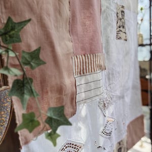 Puede incluir: Textil de patchwork con tonos rosas y blancos. Algunas secciones tienen rayas o patrones, otras incluyen inserciones de encaje. El textil está colgado, con hojas verdes de hiedra en primer plano. Estilo vintage.